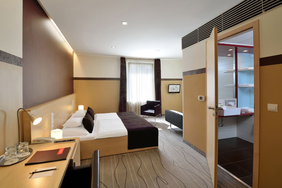 Hotellihuone, jossa on sänky, yöpöytä, lamppu, tuoli ja näkymä kylpyhuoneeseen.