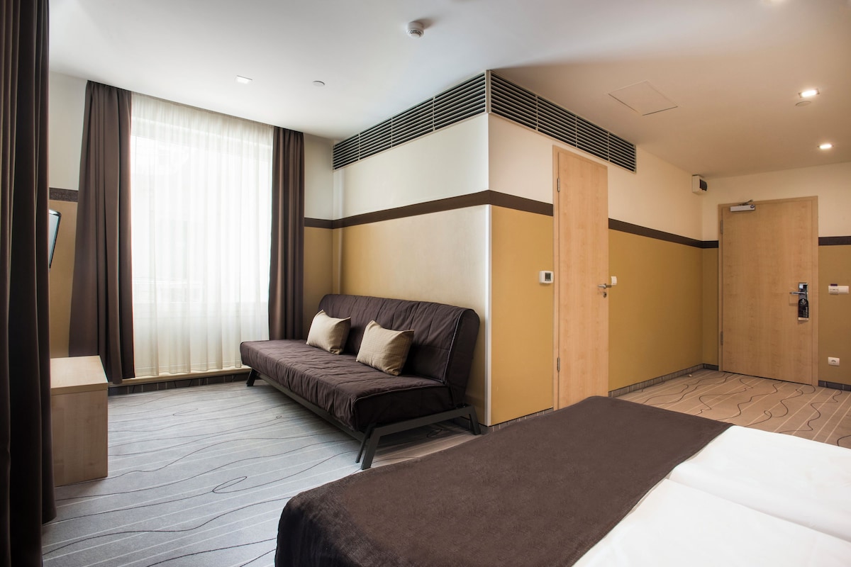 A hotel room with a bed, a sofa, a wooden wardrobe, and a window with curtains.