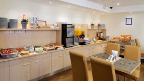 Un moderno comedor para desayunos con barra libre, que incluye dispensador de zumos, diversas opciones de comida y disposición de mesas y sillas.