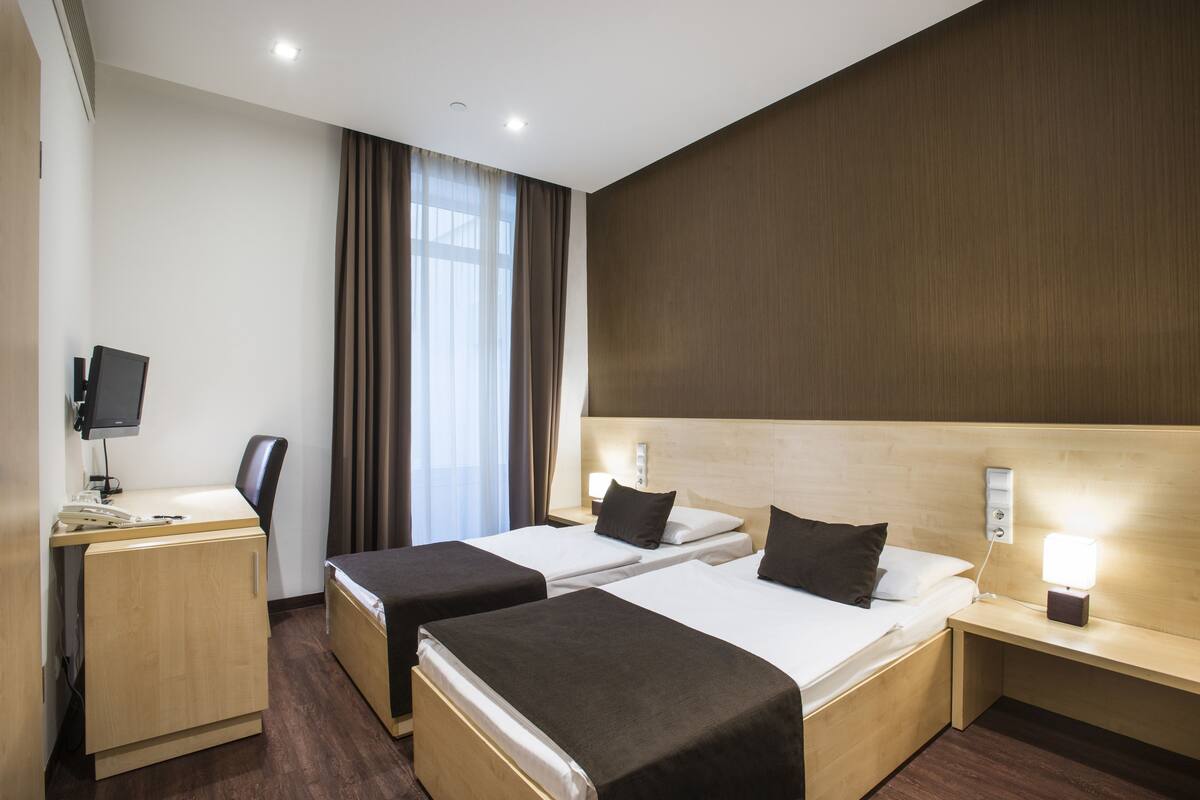 Hotellihuone, jossa on kaksi sänkyä, työpöytä tietokoneella, televisio ja ikkuna, jossa on verhot.