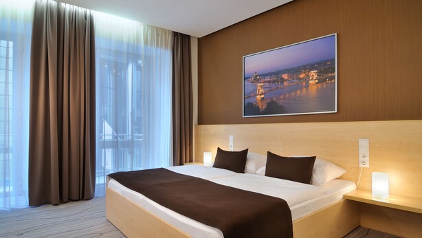 Habitación de hotel con una cama grande, una mesita de noche con lámpara, vista a un paisaje urbano a través de cortinas translúcidas y un cuadro en la pared.