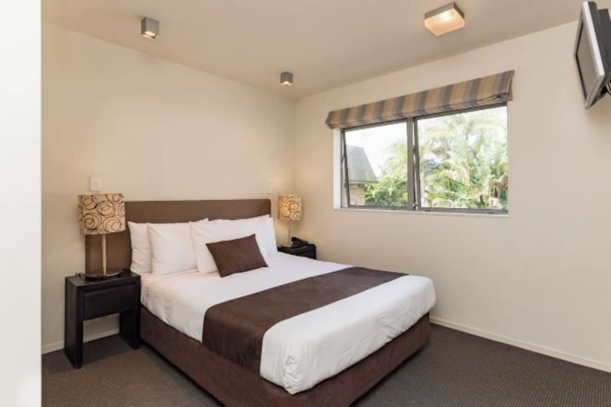 A neatly made bed with a brown headboard, two bedside lamps, and a window with a view of greenery.