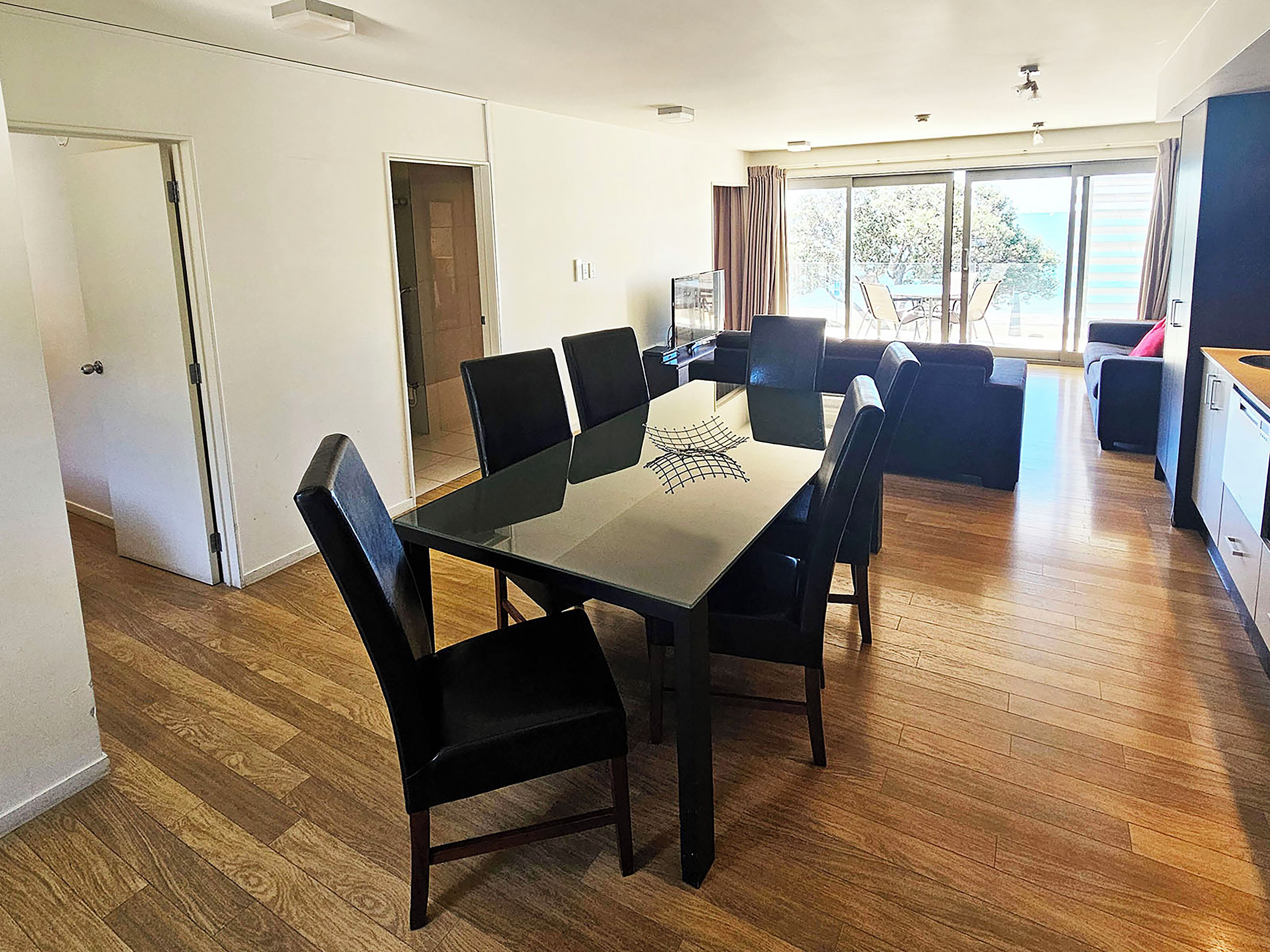 A modern dining area with a glass-top table and black chairs, a sofa, and a view of the outdoors through large windows.