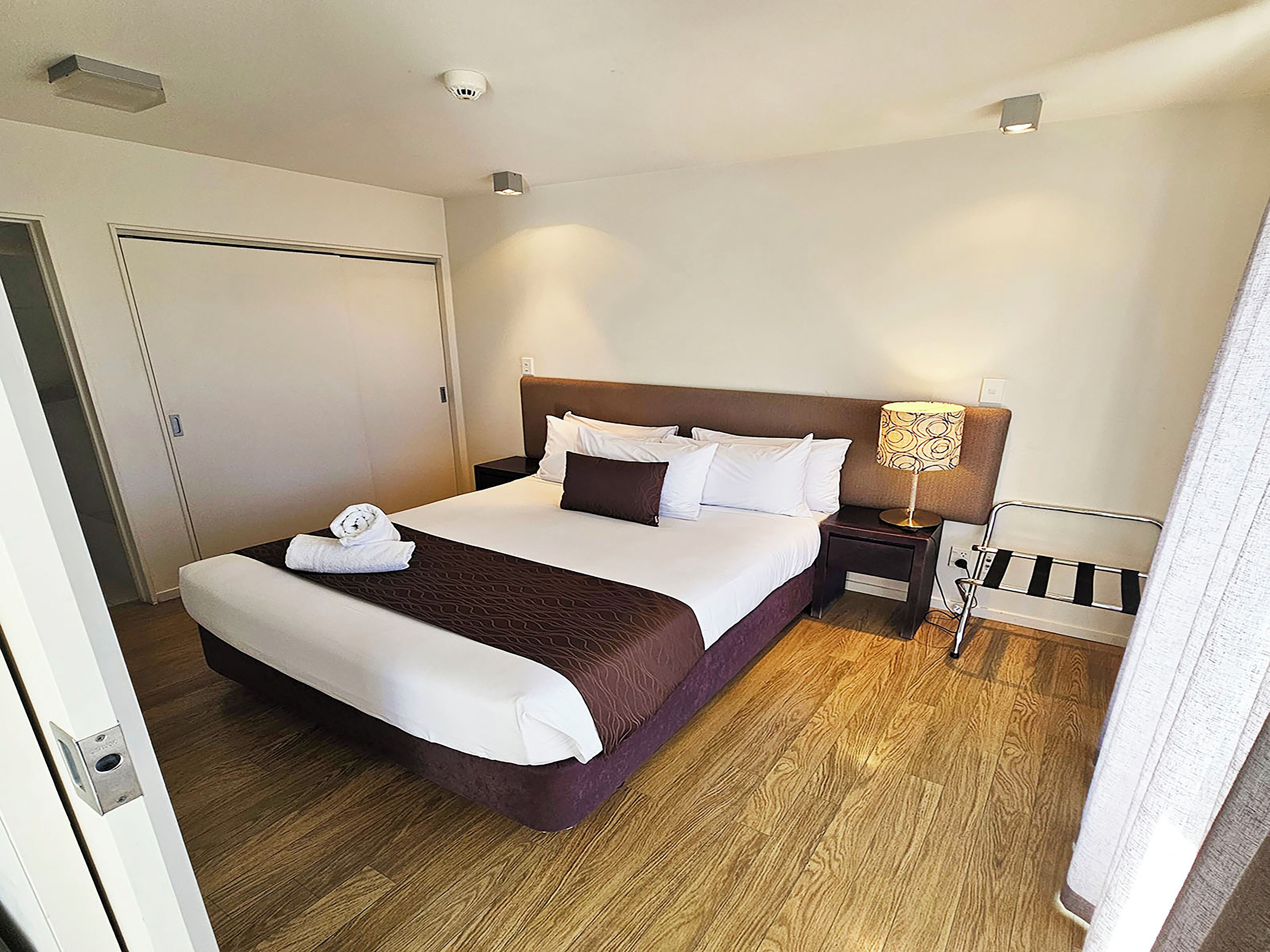 Een hotelkamer met een groot bed, een nachtkastje met een lamp, een houten vloer en een deur.