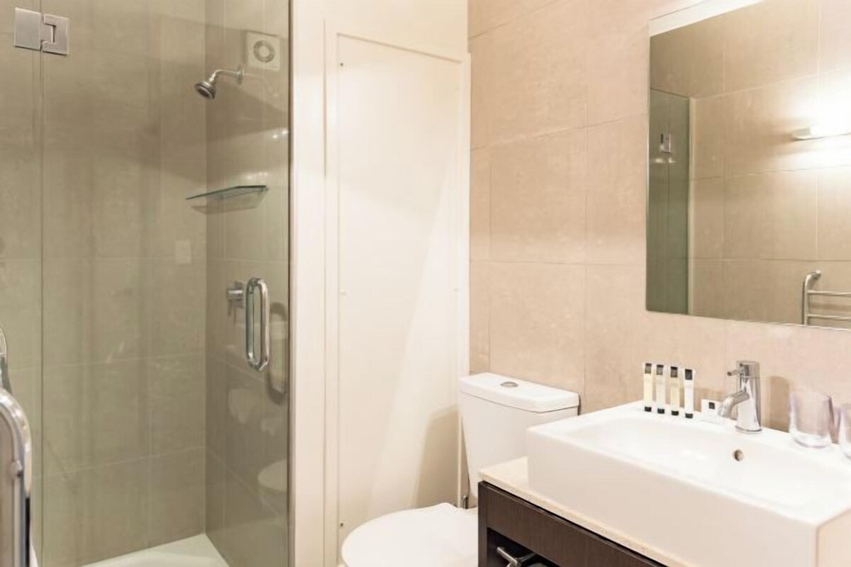 Un baño moderno con cabina de ducha de vidrio, un lavabo blanco con artículos de aseo y un espejo grande.