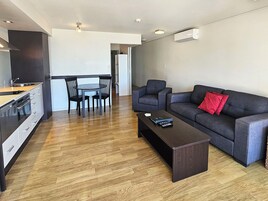 Una sala moderna con un sofá gris oscuro, una mesa de centro de madera, y un comedor con una mesa redonda oscura y sillas.
