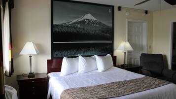 A hotel room with a bed, two bedside lamps, a ceiling fan, a large framed picture of a mountain, and a chair.