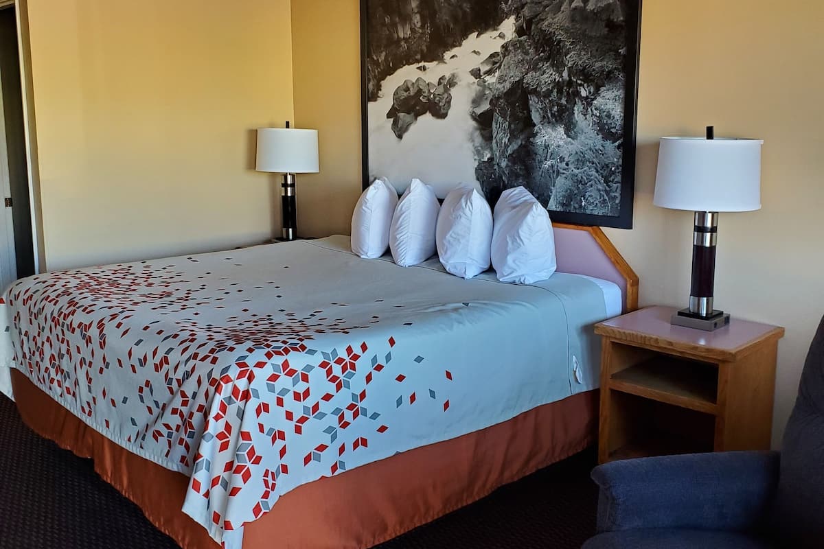A hotel room with a bed, two bedside lamps, a large painting on the wall, and a chair.