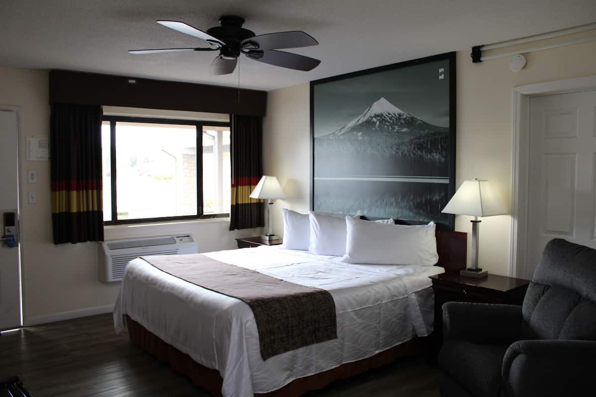 A hotel room with a bed, two bedside lamps, a ceiling fan, a large window with curtains, and a wall-mounted air conditioner.
