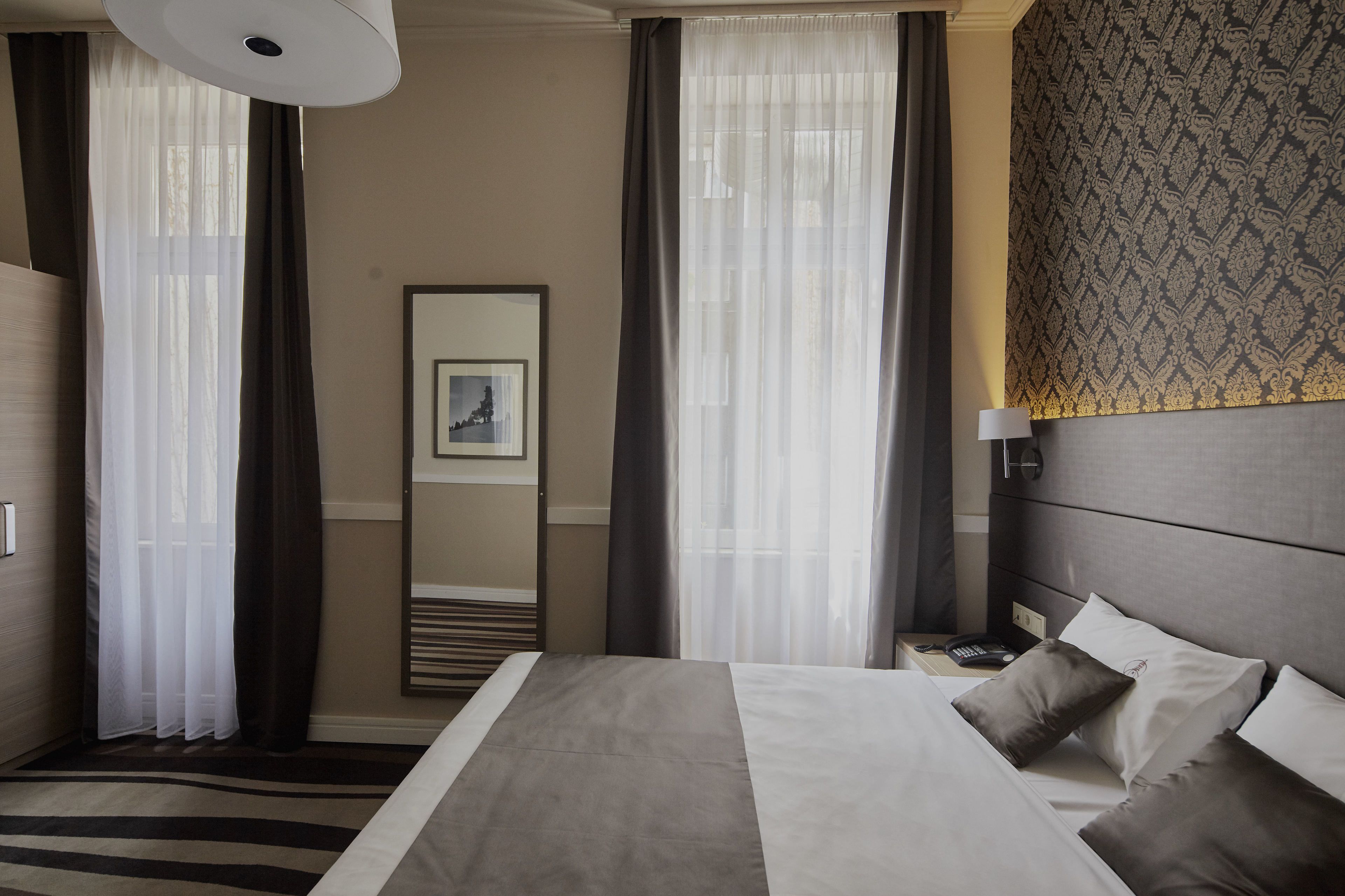 Una habitación de hotel con cama, mesita de noche, lámpara, espejo y cortinas translúcidas.