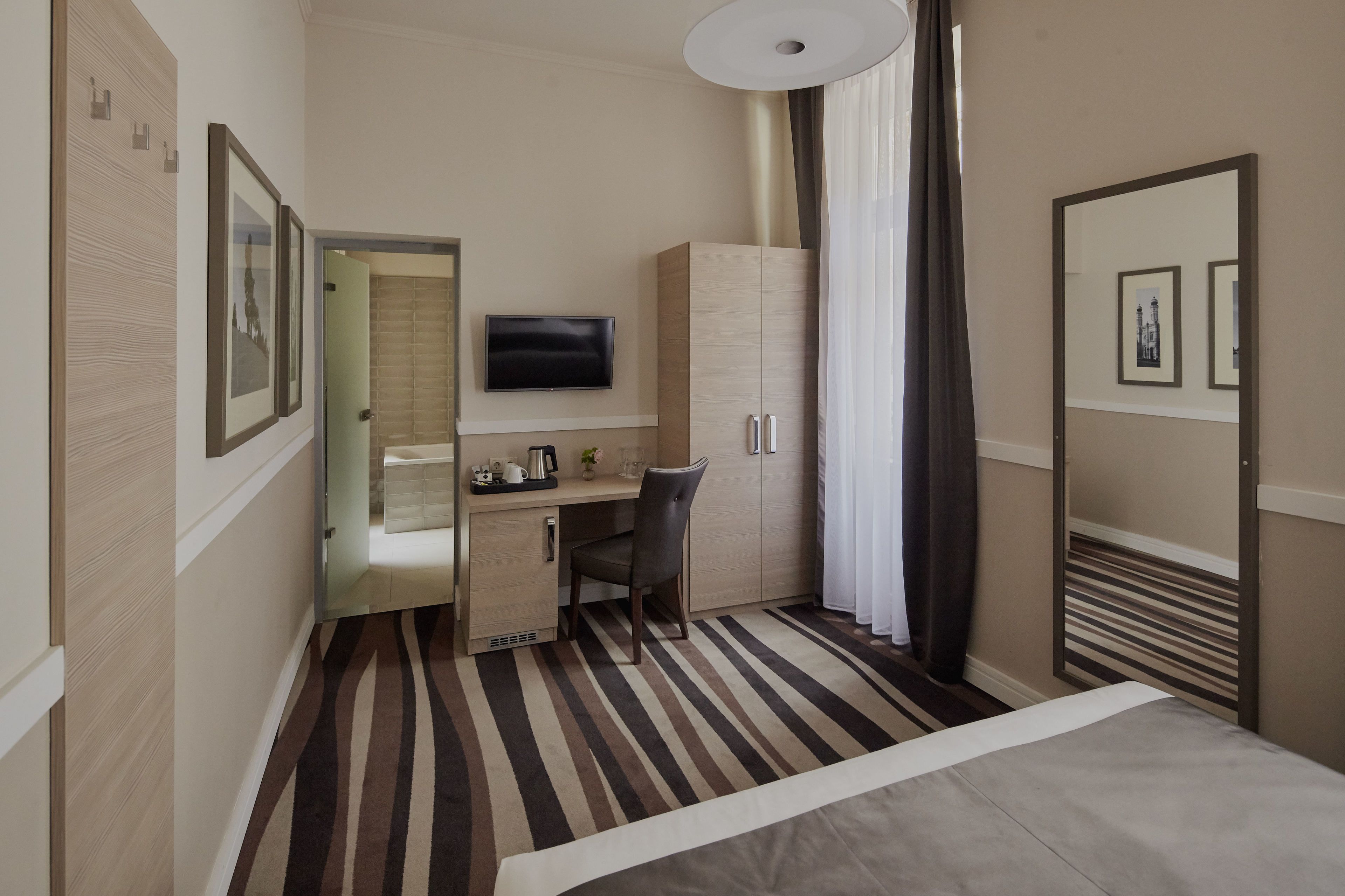 A hotel room with a bed, a desk with a chair, a television, a wardrobe, and a window with curtains.