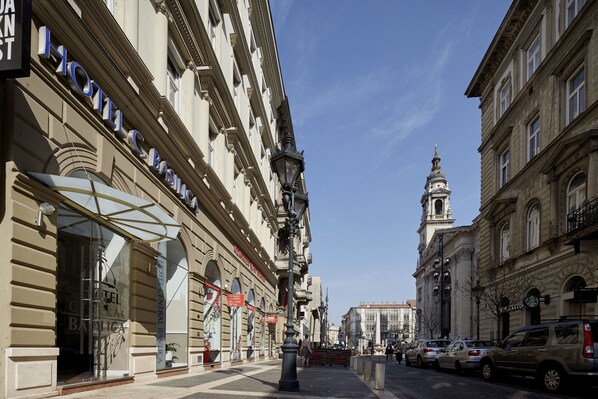 Fachada de hotel com uma grande entrada em arco, uma placa de destaque e uma torre do relógio ao fundo.