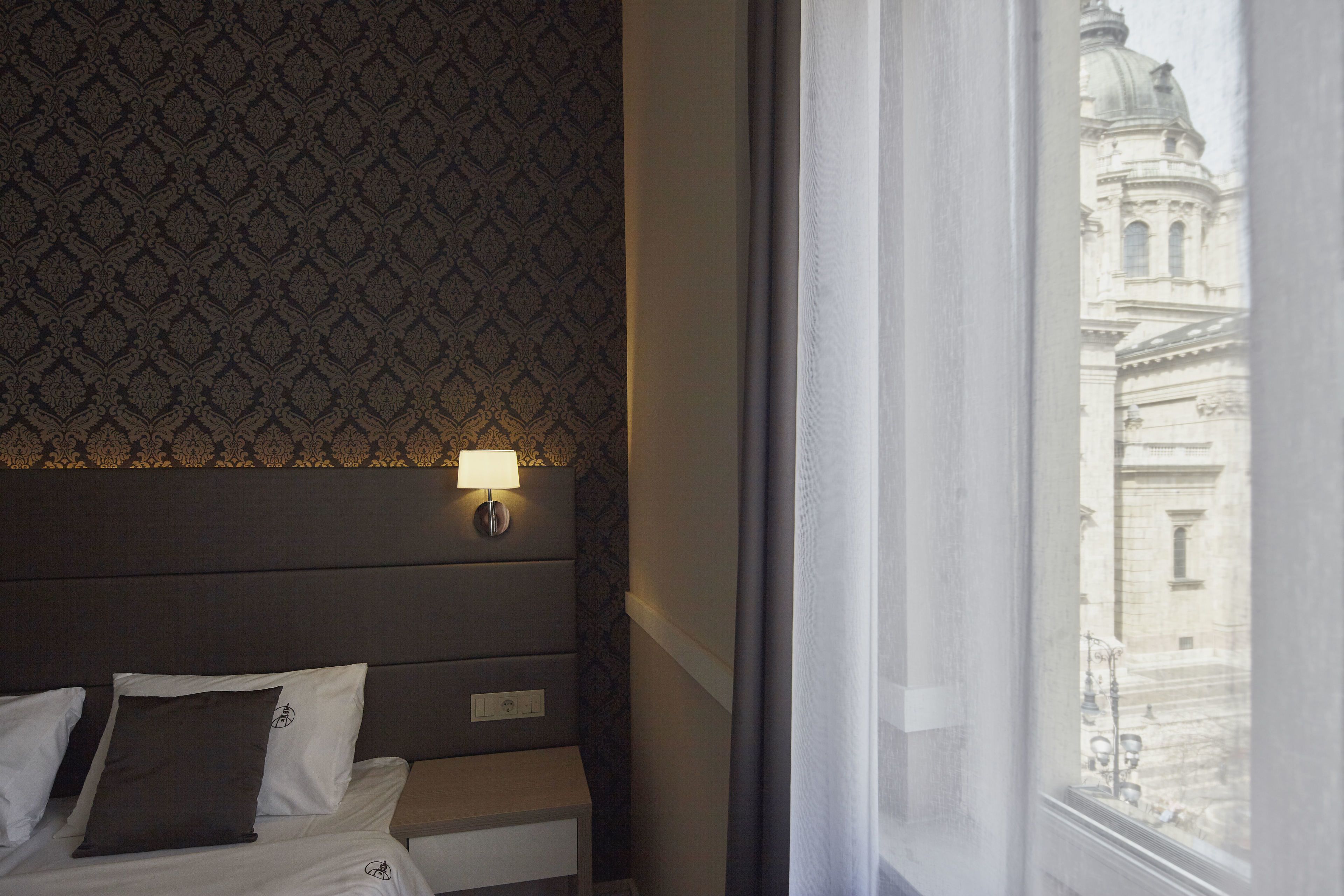 A hotel room with a patterned wallpaper, a bedside lamp, a bed with white linens, and a view of a historic building through a window.
