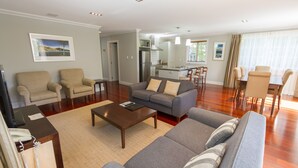 A spacious living room with a grey sofa, beige armchairs, and a wooden coffee table. A kitchen with a dining area is visible in the background. - Russell Cottages Tanoa Hotels (Russell)