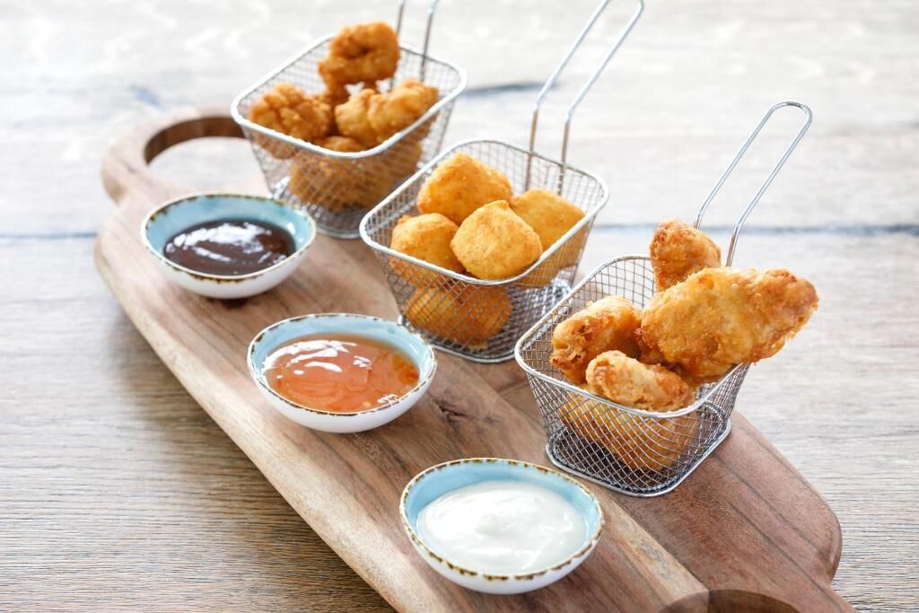 A wooden board with three wire baskets containing fried food items, accompanied by small bowls of dipping sauces.