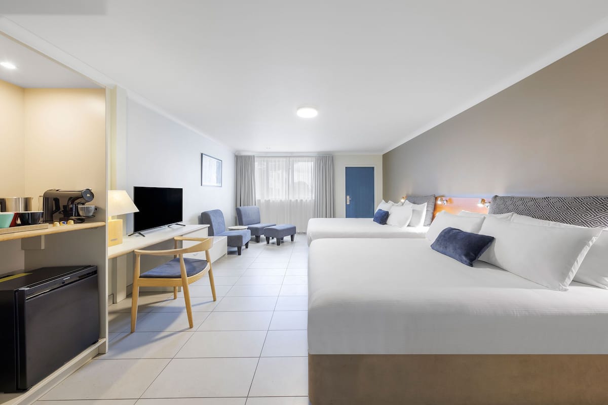 A modern hotel room with a kitchenette, a flat-screen TV, a sofa, a dining area, and a large window with curtains.