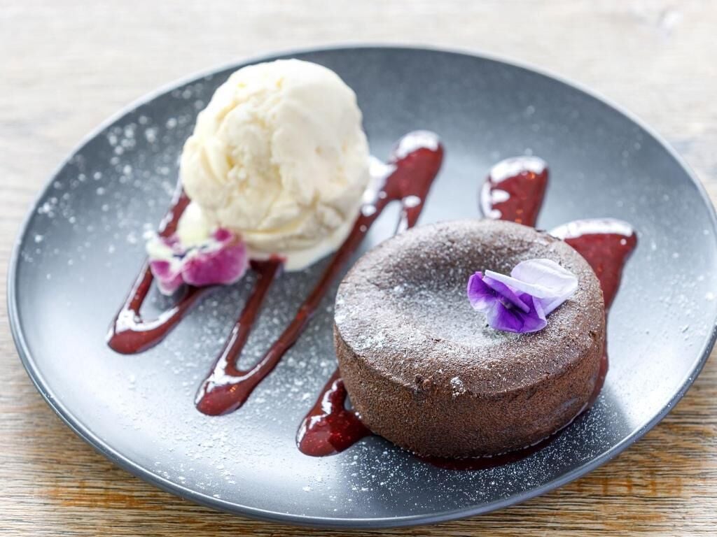 Un plato de postre con pastel de chocolate y una bola de helado de vainilla, decorado con una flor morada y bañado en salsa roja.