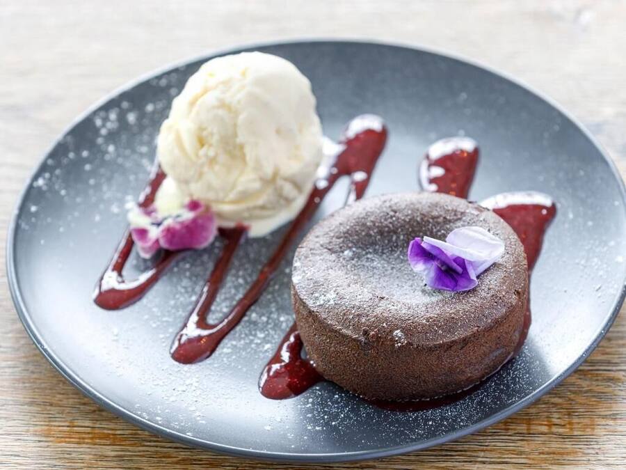 Un plato de postre con pastel de chocolate y una bola de helado de vainilla, decorado con una flor morada y bañado en salsa roja.