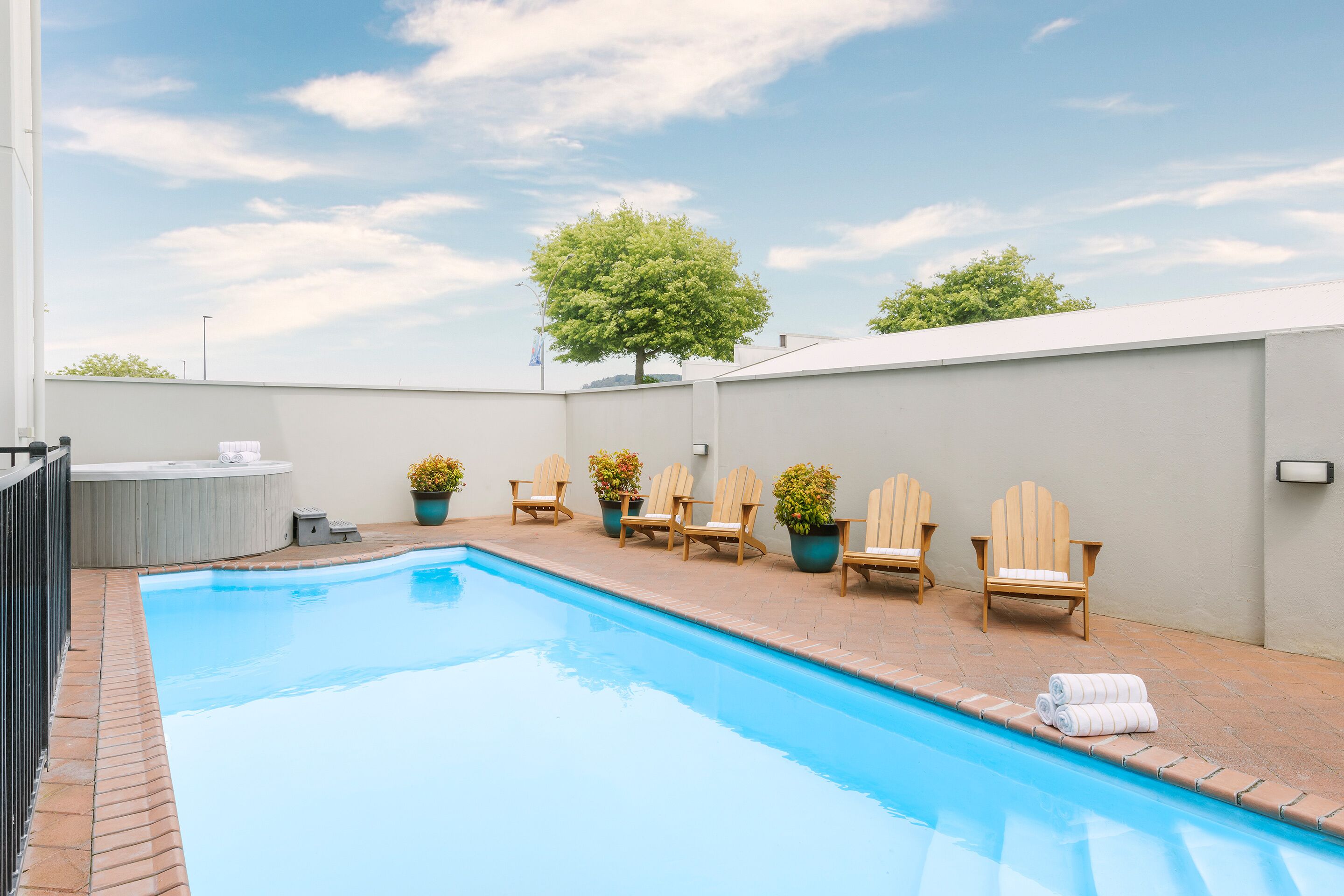 A swimming pool with six Adirondack chairs and potted plants, a hot tub, and a tree in the background.