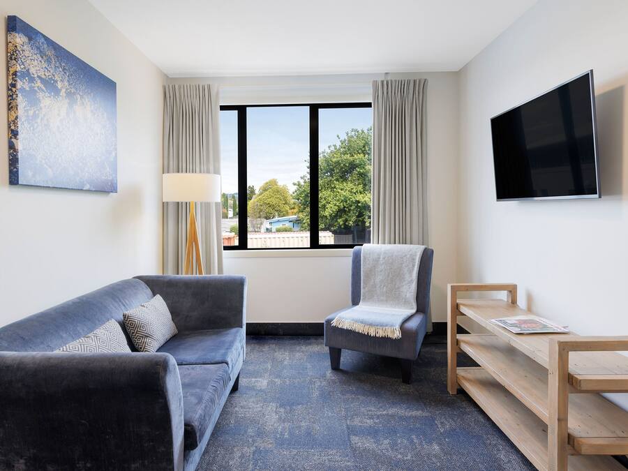 Una habitación de hotel moderna con sofá, sillón, banco de madera y televisor montado en la pared.