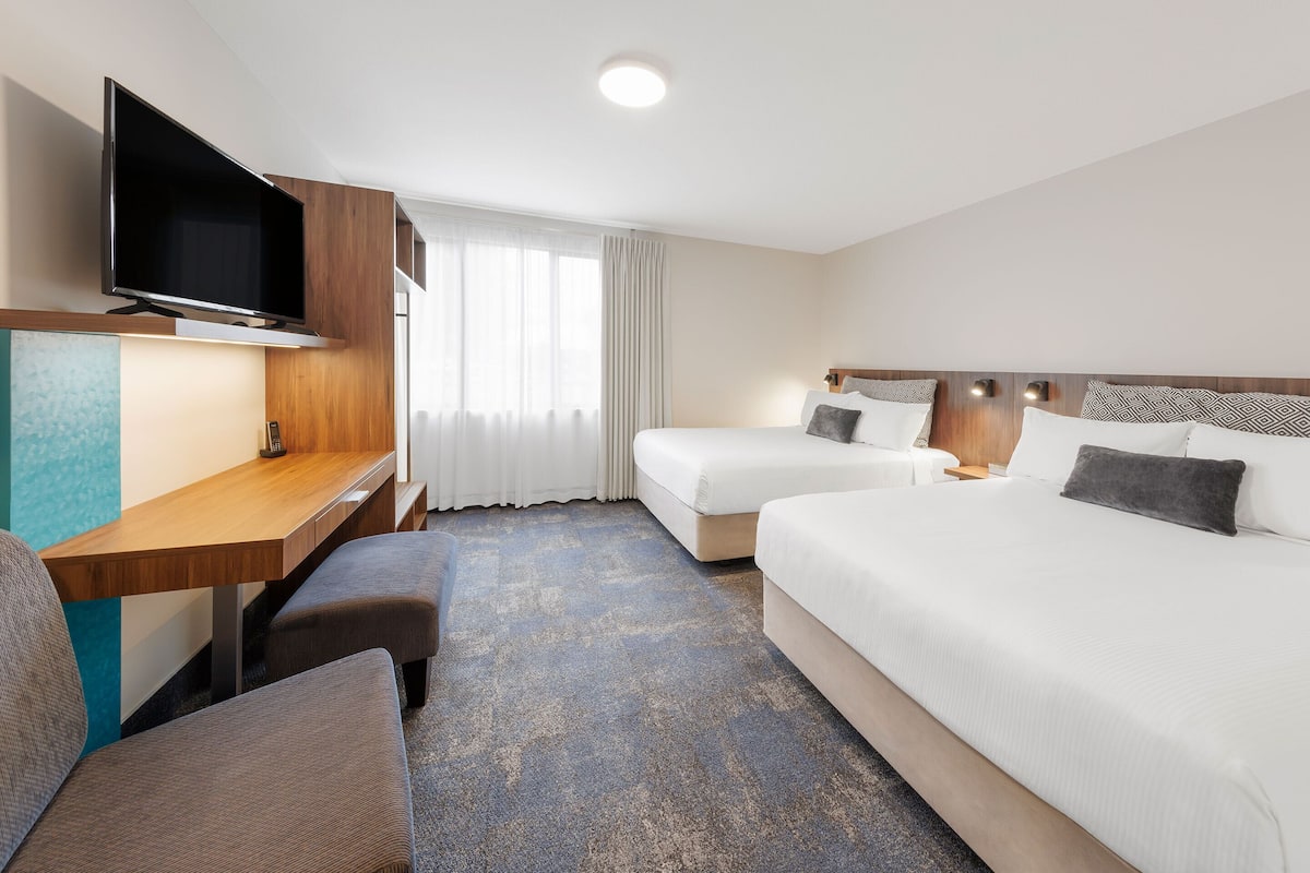 A hotel room with a flat-screen TV mounted on the wall, a desk with a chair, two beds with pillows, and a large window with curtains.