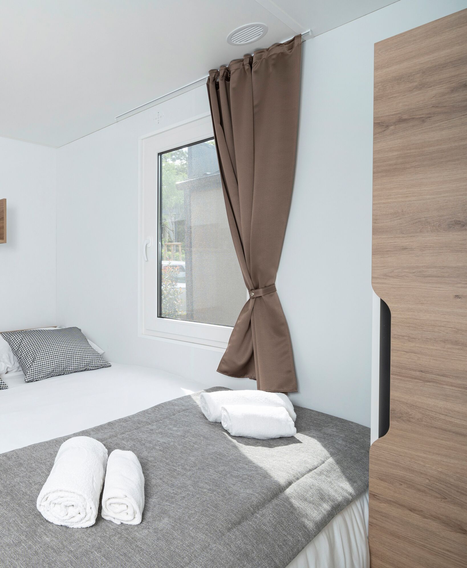 Un dormitorio moderno con cama, manta gris, dos toallas enrolladas, ventana con cortinas y un armario de madera.