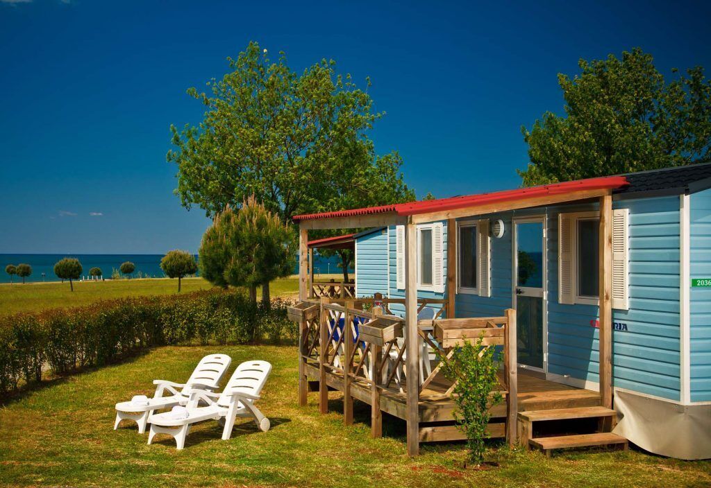Una cabaña pequeña azul con una terraza de madera, dos sillones blancos y vistas al mar.