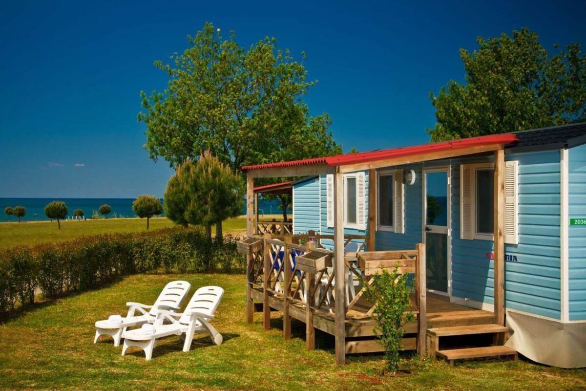 Una cabaña pequeña azul con una terraza de madera, dos sillones blancos y vistas al mar.