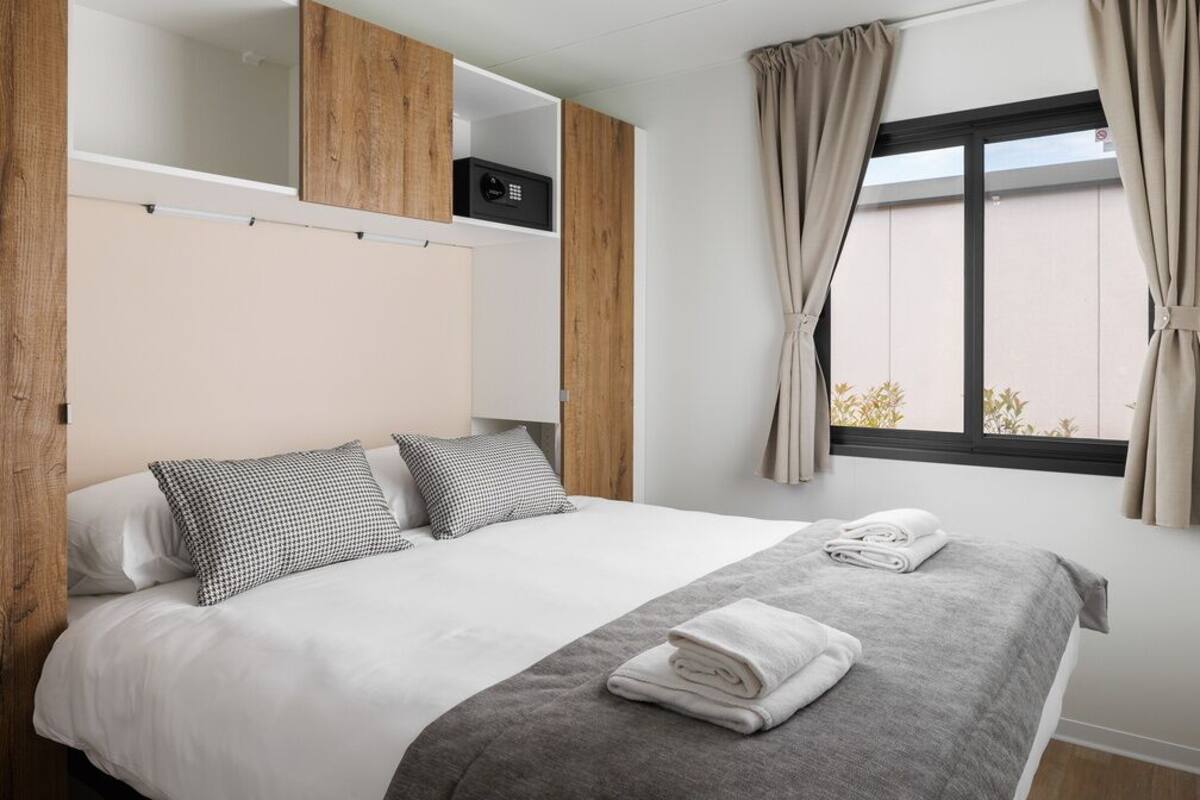 Una cama bien hecha con ropa de cama blanca y gris, un plaid gris y dos almohadas. Se observa un pequeño dispositivo negro sobre una repisa de madera.