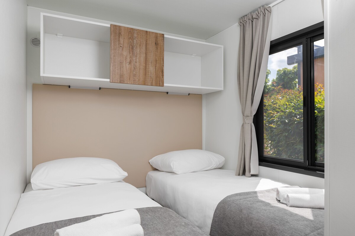 Une chambre d’hôtel avec deux lits, une étagère en bois, une fenêtre avec des rideaux et une vue sur la verdure.