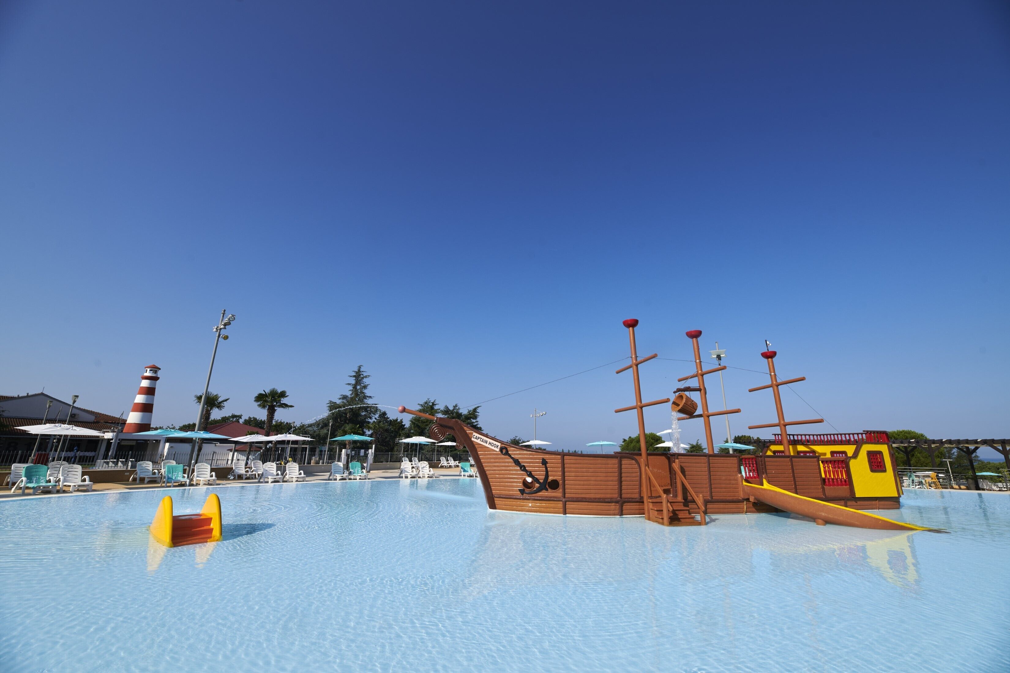 A swimming pool with a pirate ship-themed slide, lounge chairs, and a lighthouse in the background.