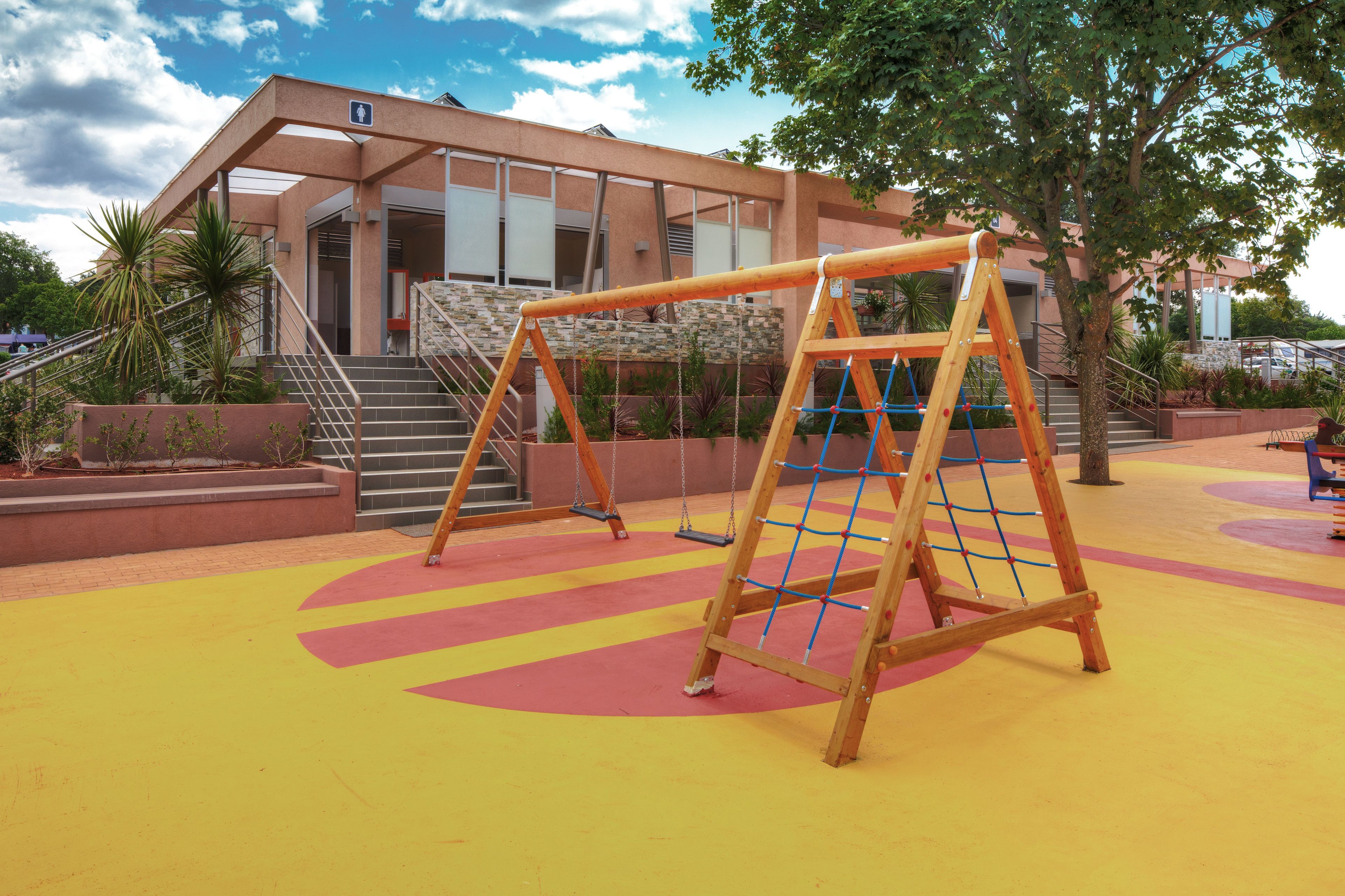 Une aire de jeux équipée de balançoires et d’un filet d’escalade, entourée d’un bâtiment doté d’escaliers et d’une rampe d’accès pour les personnes à mobilité réduite.