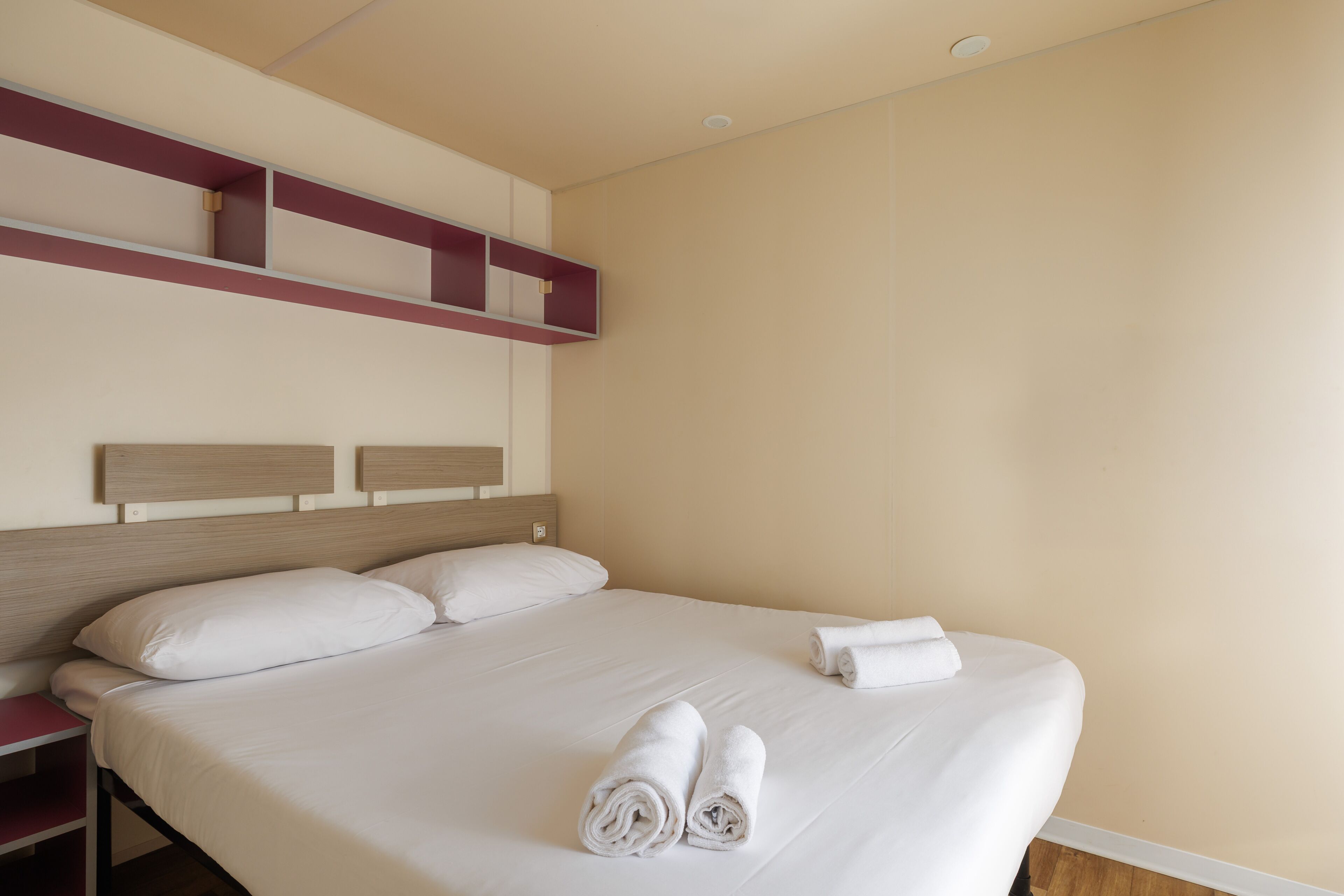 A hotel room with a large bed, two pillows, a wooden headboard, a red shelf, and two rolled towels on the bed.