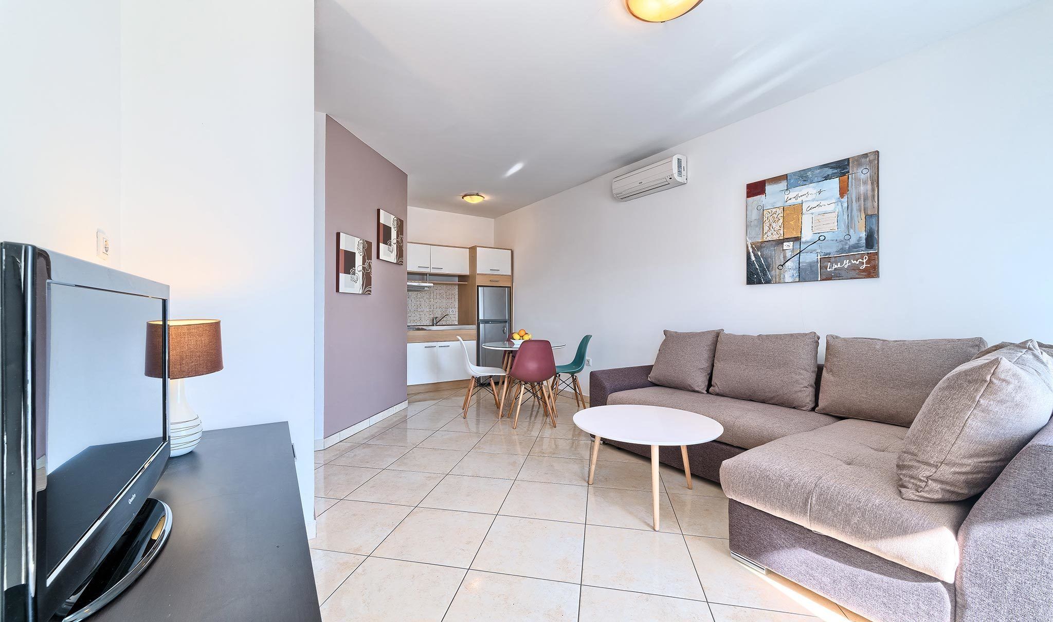 Uma sala de estar moderna com televisão de tela plana, um sofá, uma mesa de centro e um espaço de jantar com cadeiras e uma pequena mesa.
