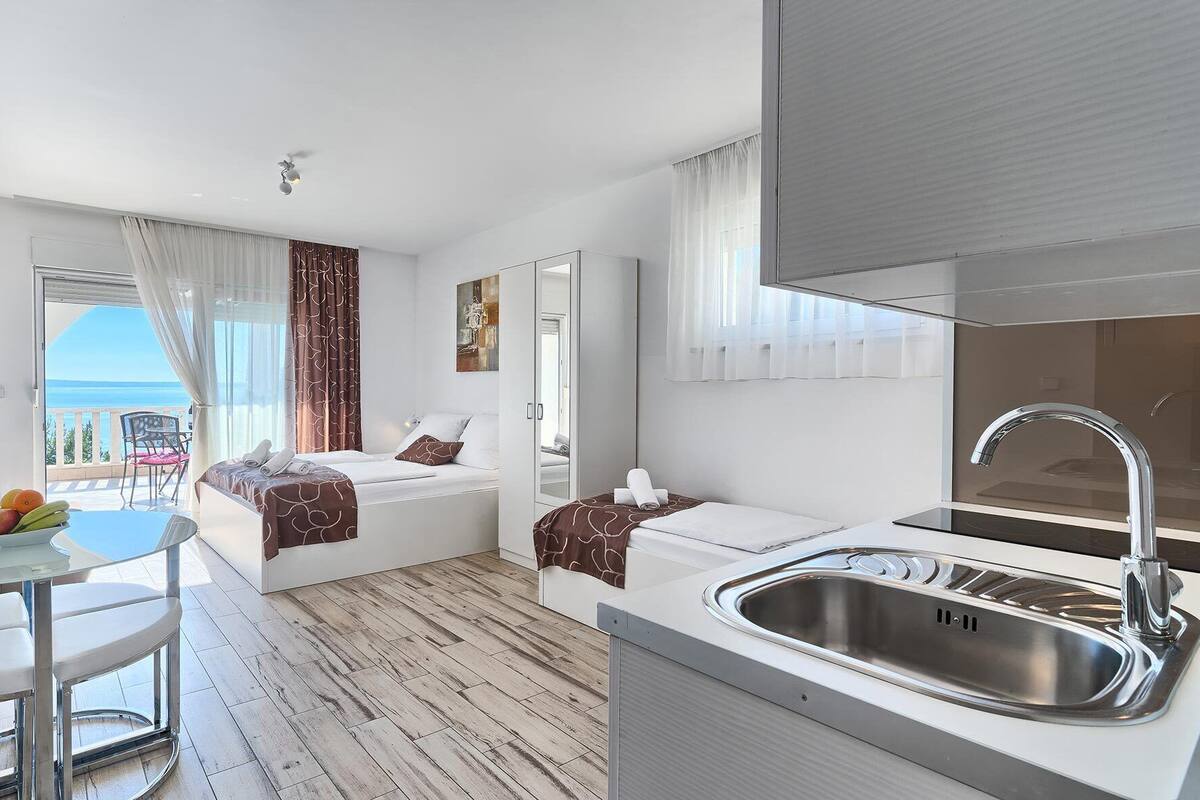 A modern hotel room with a balcony, a bed, a small table with chairs, and a kitchen area with a sink.