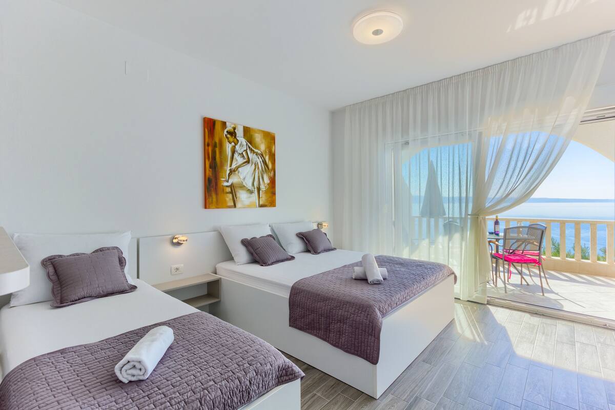 A modern hotel room with two beds, a painting on the wall, and a balcony with a view of the sea.