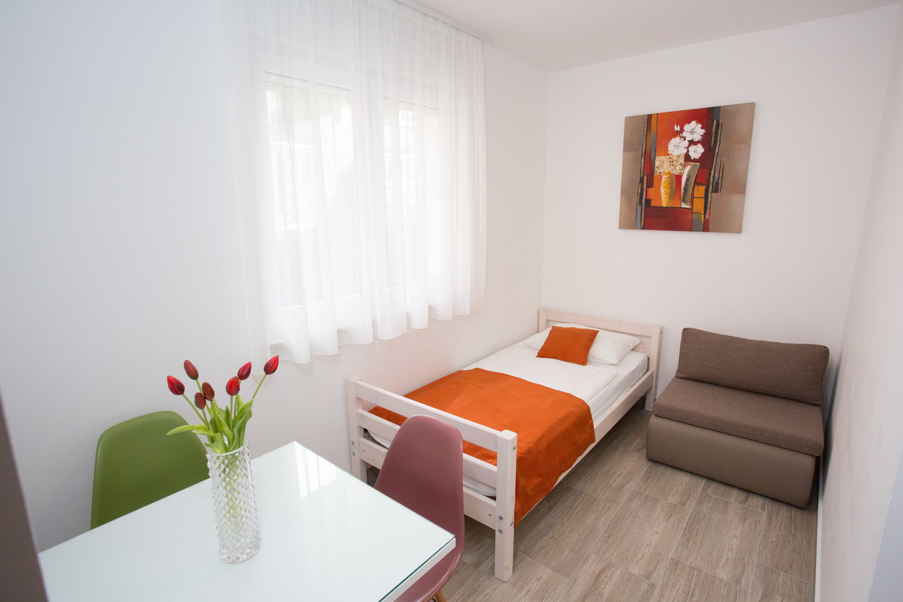 Ein kleiner, hell eingerichteter Raum mit einem Bett, einem Schlafsofa, einem Tisch und einem Stuhl. Es gibt ein Fenster mit durchsichtigen Vorhängen, ein Bild an der Wand und einen Blumenstrauß auf dem Tisch.