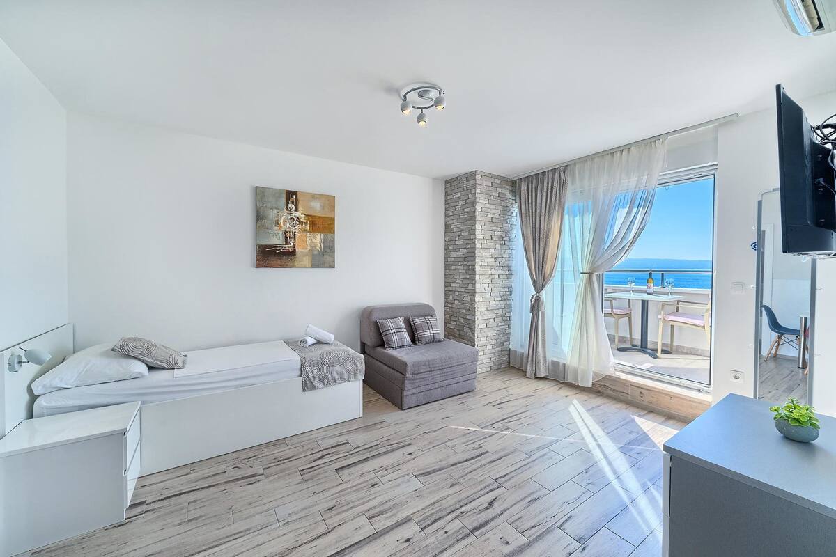 A modern hotel room with a bed, a seating area with a view, a stone accent wall, and a ceiling light.