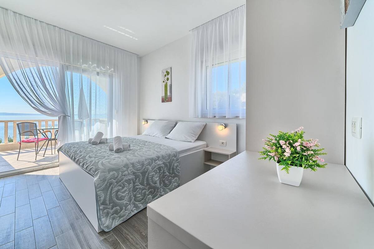 A bedroom with a bed, a bedside table, a potted plant, and a view of the sea through a balcony.