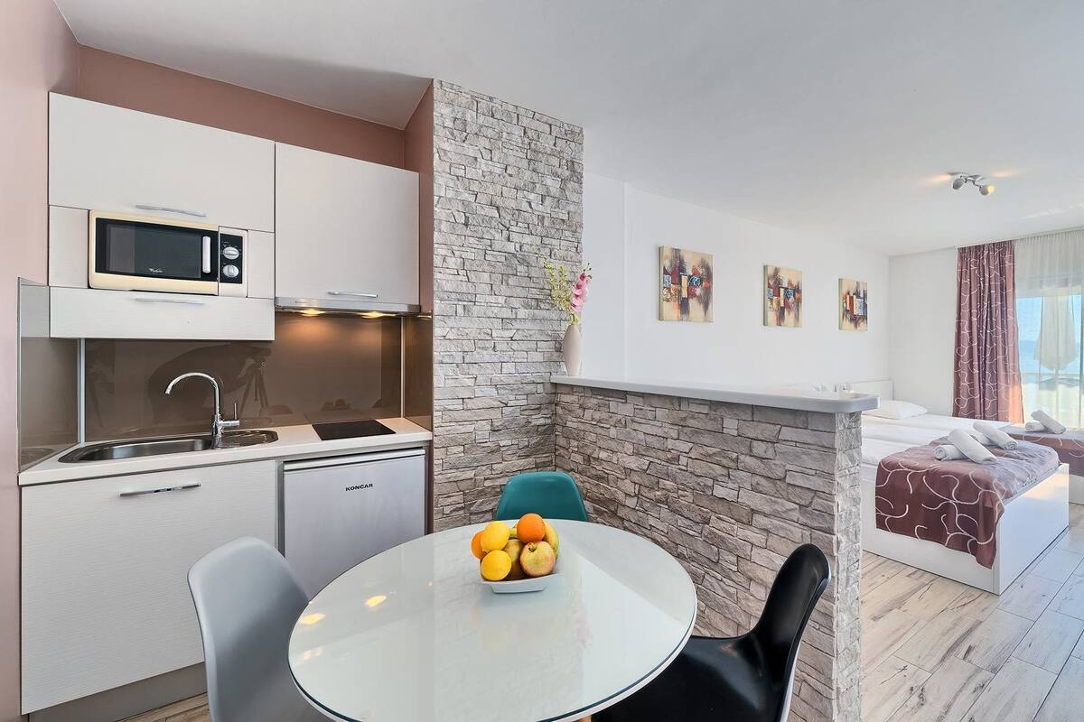A modern kitchen with a microwave, sink, and a dining area with a table and chairs. There is a stone wall, a bed with a view, and artwork on the wall.