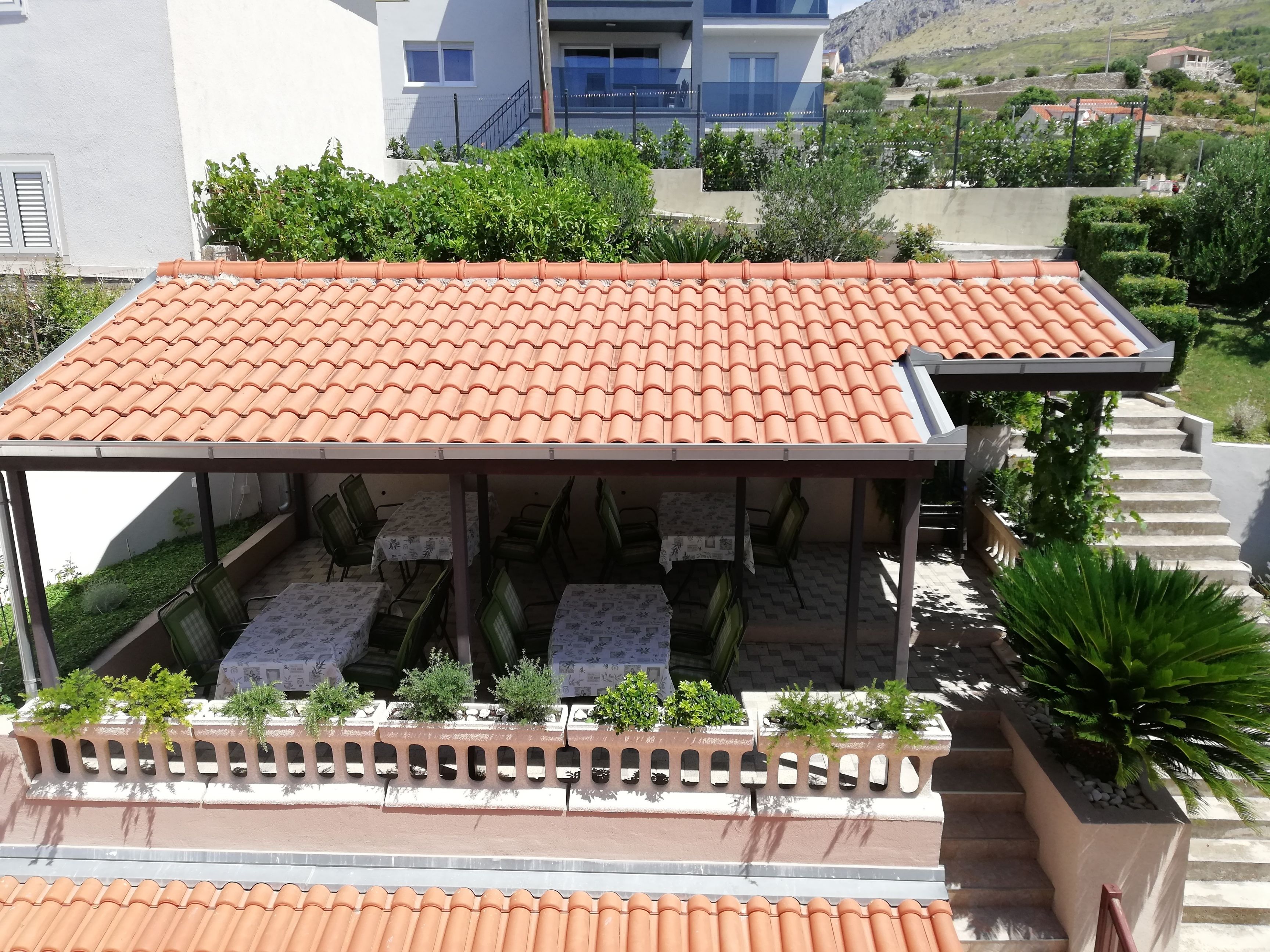A covered outdoor seating area with tables and chairs, surrounded by greenery and a building in the background.