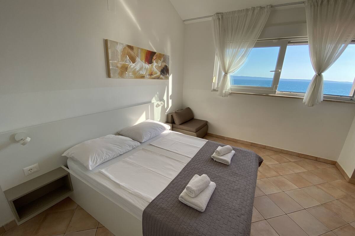 A hotel room with a bed, a window with curtains, a painting on the wall, and a small seating area.