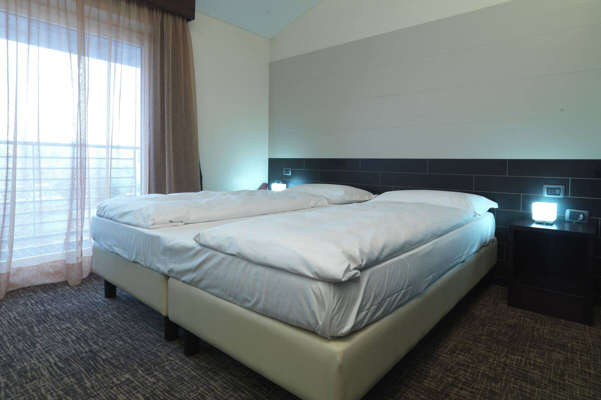 Habitación de hotel con cama doble, mesita de noche con lámpara y ventana con cortinas translúcidas.
