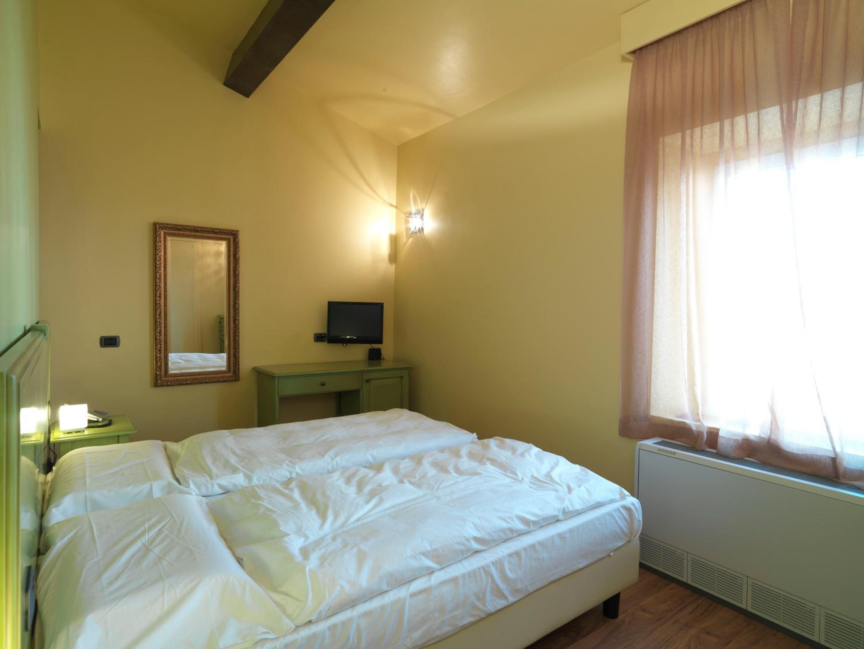 A bedroom with a bed, a desk, a television, a mirror, and a window with curtains.