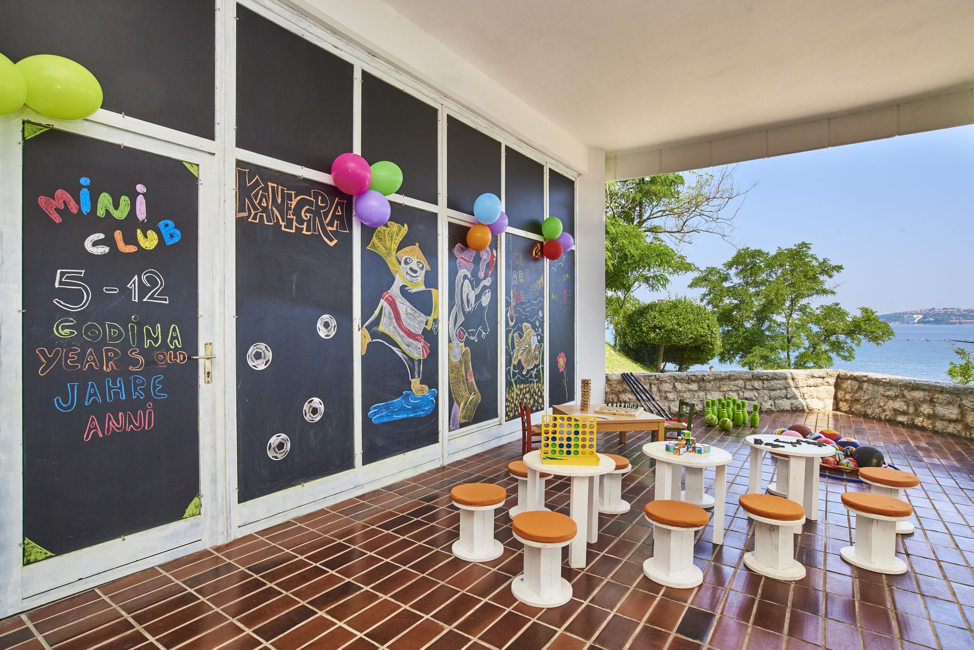 Zona infantil con cartel de pizarra, asientos exteriores y vista al mar.