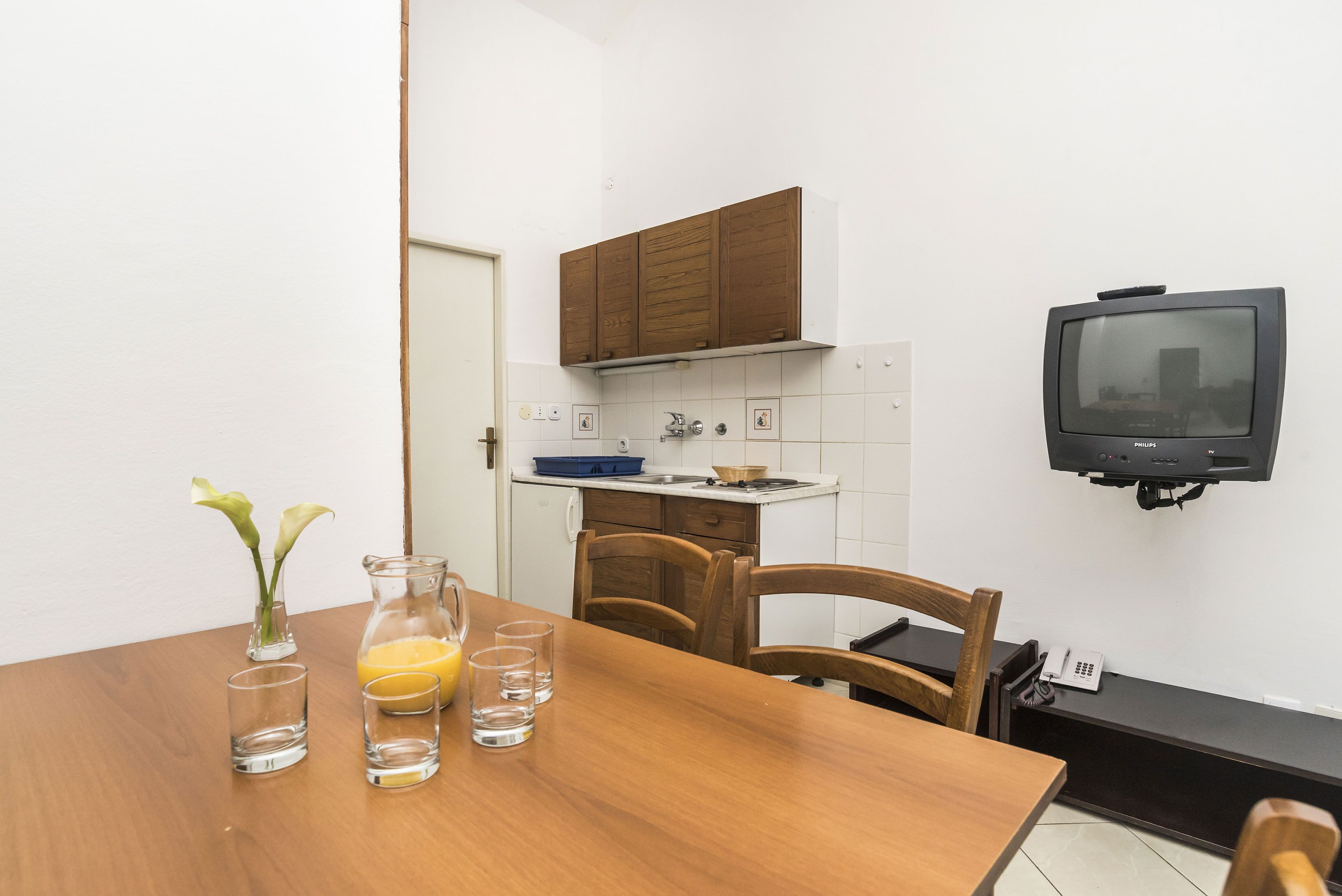 A compact kitchen with a wooden cabinet, a small table with a vase and glasses, and a television mounted on the wall.