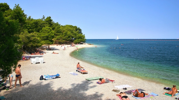Una playa con gente tomando el sol, un velero a lo lejos y un cielo azul despejado.