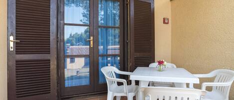 Une terrasse avec une table et des chaises blanches, une porte vitrée avec des volets et un feu rouge d’urgence.