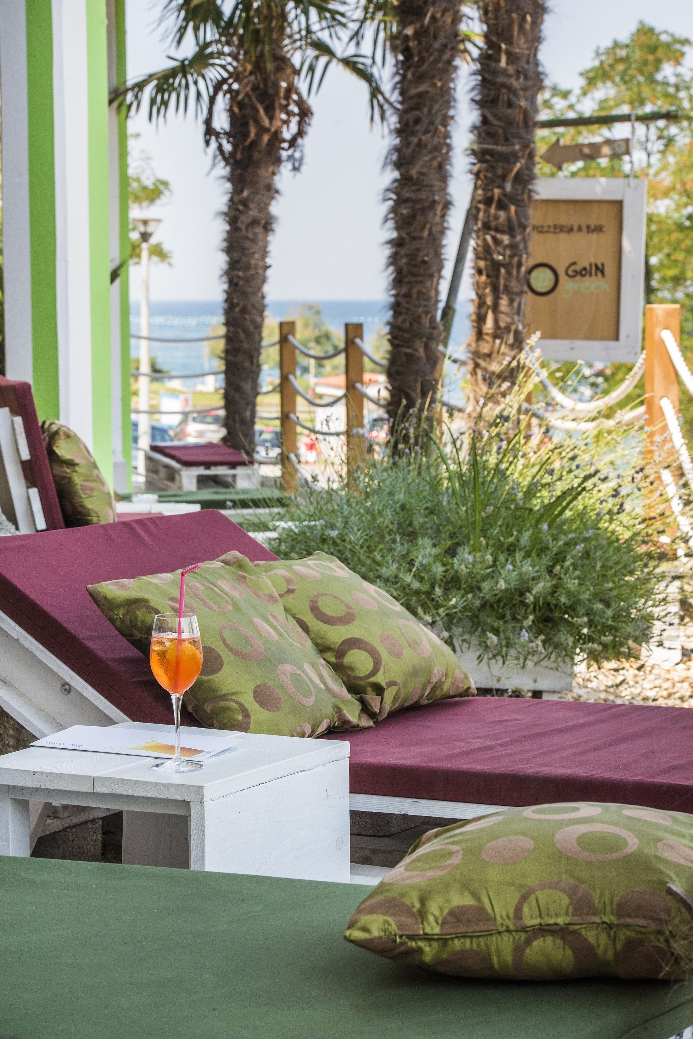 Un salon de plage avec un verre d’une boisson orange, des coussins aux motifs verts et bruns, et une pancarte indiquant « Goin Green ».