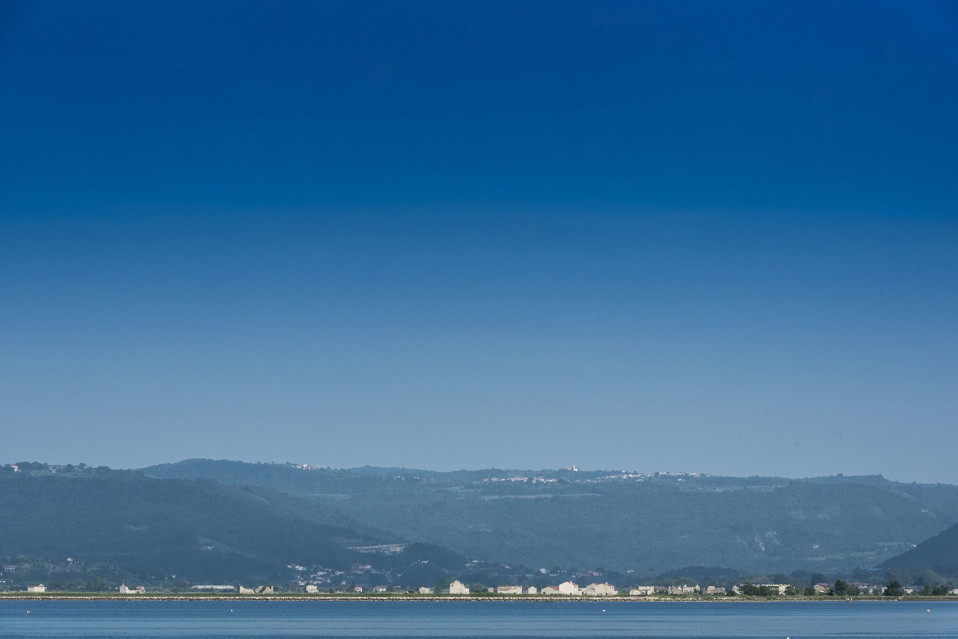 A coastal view with a clear sky, a body of water, and a distant hill with buildings.